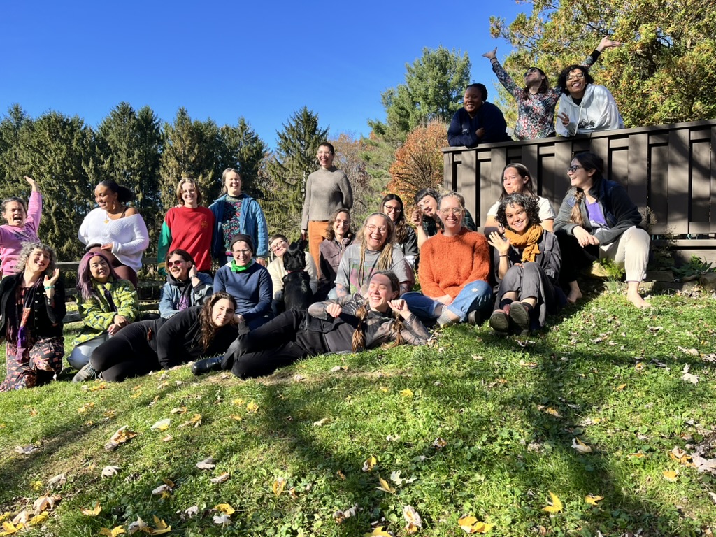 A photograph of many herb students with big smiles, sitting, laying and standing in dappled autumn sunlight, blissed out on plant vibes and exuding pure love.